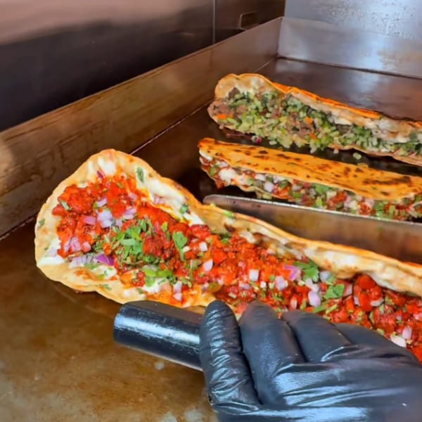 A chef's gloved hand prepares large, vibrant machete tacos filled with meat, onions, and cilantro on a griddle.