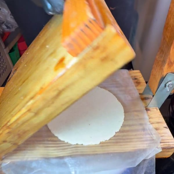 A round, white tortilla being pressed flat by a wooden tortilla press.